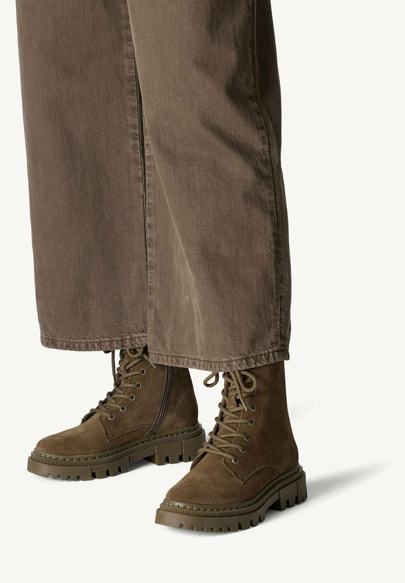 Bottes de combat en daim marron à lacets avec fermeture éclair latérale et semelles épaisses en caoutchouc, portées avec un pantalon marron à jambes larges.