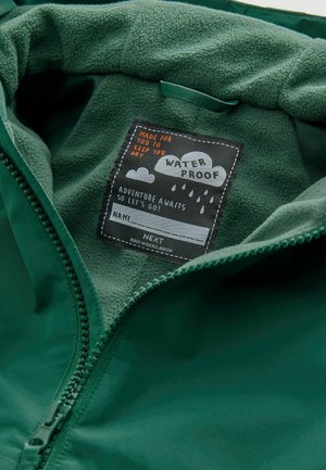 Wodoodporna zielona kurtka ma miękką fleecową podszewkę oraz czarną metkę z białym i pomarańczowym tekstem. Zamek błyskawiczny.