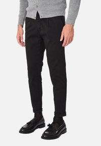 Pantaloni neri a gamba stretta con orli a fondo cavo, realizzati in un tessuto morbido, abbinati a mocassini neri lucidi e a un cardigan grigio chiaro.