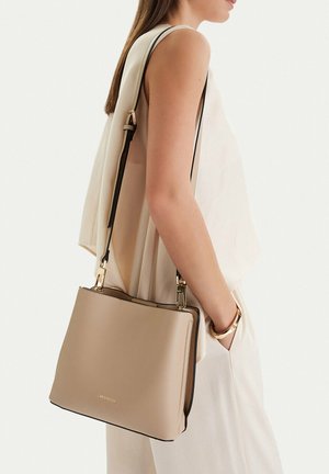 Žena v béžovém topu bez rukávů a kalhotách nese přes rameno světle hnědou kabelku se zlatým kováním a černým popruhem.