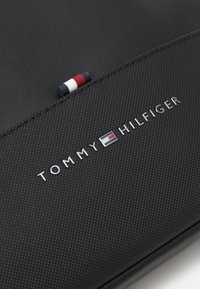 Borsa in pelle nera con texture, caratterizzata da un logo argentato "TOMMY HILFIGER" e un piccolo accento rosso, bianco e blu vicino alla cerniera.