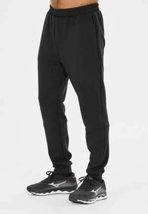 Man wearing black athletic jogger pants and black running shoes with white soles, standing against a plain light background.