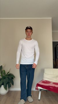 Camisa branca de mangas longas com mangas ajustadas, jeans azuis, chinelos cinzentos e um boné preto com um logótipo, de pé contra uma parede lisa.