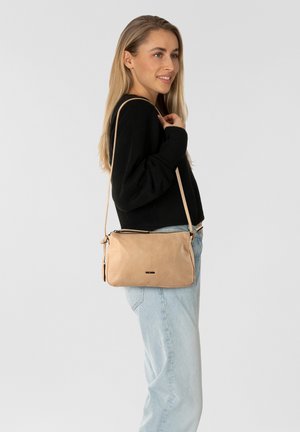 Borsa a tracolla in pelle beige con finitura testurizzata, chiusura con zip e tracolla regolabile. Modello che indossa un maglione nero e jeans chiari.