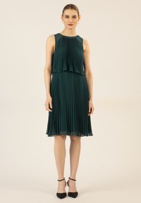 Grün plissiertes Kleid mit ärmellosem Design, geschichteten Oberteil und mittellanger Rocklänge. Getragen mit schwarzen spitzen Pumps, die eine glatte Textur zeigen.