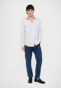 Camisa abotonada con cuello, de rayas verticales azules y blancas, combinada con jeans azules y mocasines negros. La modelo está de pie frente a un fondo liso.