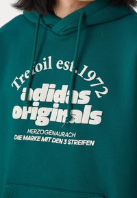Grön bomullshoodie med dragsko, prydd med kraftig vit text: "Trefoil est. 1972," "adidas originals," och ytterligare varumärkesdetaljer.