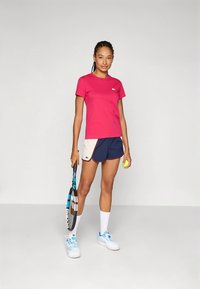 T-shirt rosa de manga curta, calções azul-marinho e creme com detalhes verdes, segurando uma raquete e uma bola de ténis, complementado com meias e sapatilhas brancas.