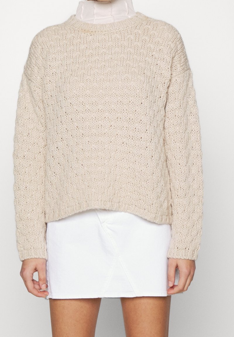 Pull en maille épaisse beige avec motif torsadé, associé à une mini-jupe blanche portée par une personne, montrant le torse et le haut des jambes.