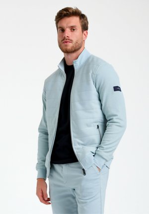 Giacca a zip blu chiaro con texture trapuntata, colletto alto e tasche laterali. Indossata sopra una maglietta nera, con un logo sul manico.