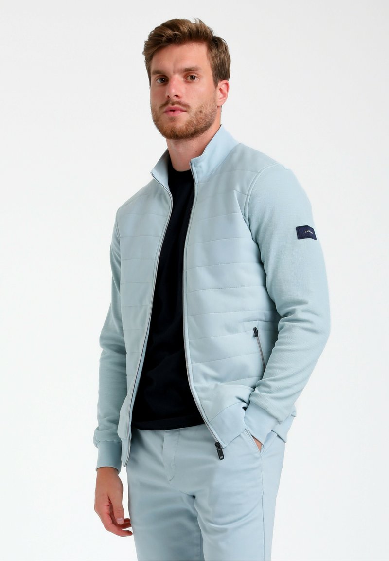 Giacca a zip blu chiaro con texture trapuntata, colletto alto e tasche laterali. Indossata sopra una maglietta nera, con un logo sul manico.