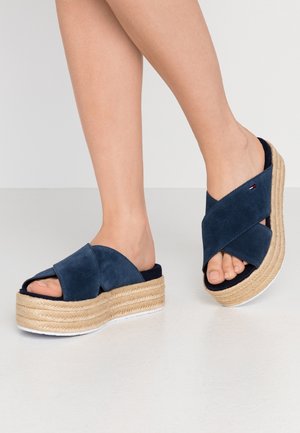 Voeten met marineblauwe suède plateausandalen met gekruiste bandjes en zolen van gevlochten touw tegen een neutrale achtergrond.