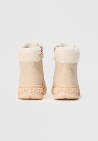 Beige ankelstøvler med fluffy mansjetter, sidelukking med glidelås, og preget "Tommy Hilfiger" merkevare på gummisålene, vist fra baksiden.