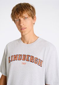 Grå T-shirt med kort ärm som har "LINDBERGH 1927" broderat i orange och svart. Enkel design med rundad halsringning, mjuk textur.