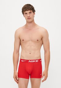 Ung mand står mod en hvid baggrund iført røde Nike boxershorts med hvidt logo på taljen og benet.