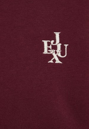 Burgundska pamučna tkanina s bijelim izvezenim slovima "E", "J", "U" i "X" u preklapajućem dizajnu s mekanom, teksturiranom površinom.