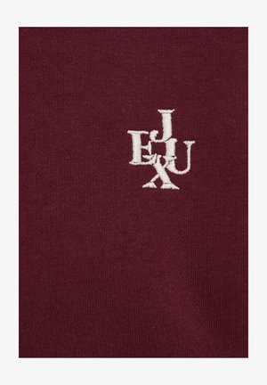 Bordo kokvilnas audums ar baltu izšūtu burtiem "E", "J", "U" un "X", kas veido savstarpēji pārklājas dizainu ar maigu, teksturētu virsmu.