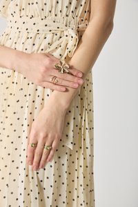 Robe crème à pois avec une taille nouée, mettant en avant des anneaux en or avec des fleurs et des motifs géométriques aux doigts. Texture notable visible dans le tissu et les bijoux.