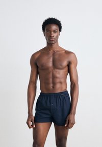 Boxers en coton bleu marine avec une taille élastique et un bouton à l'avant. Le modèle a une silhouette tonique, se tenant devant un fond clair.