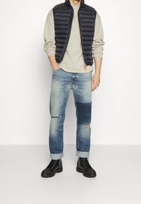 Mann in einem beigen Sweatshirt, schwarzem Steppweste, patchwork-rotblauen Jeans mit hochgekrempelten Säumen und schwarzen Schnürstiefeln, der vor einem schlichten Hintergrund steht.