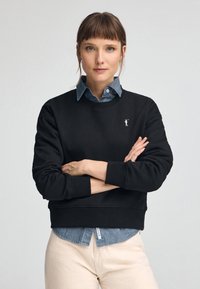 Svart sweatshirt med rund hals och ribbade ärmslut, lagd över en ljusblå denimskjorta; har en liten vit logotyp på bröstet.