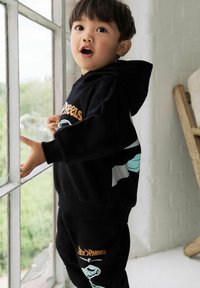 Young child in black hoodie and pants with orange and blue Hot Wheels logos standing by a window, looking outside with mouth slightly open.