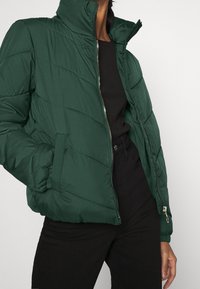 Giacca bomber verde con colletto alto, cuciture diagonali, chiusura con zip e tasche laterali. Tessuto liscio con polsini ed orlo elasticizzati.