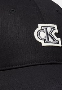 Gorra de béisbol negra con un parche bordado blanco con las letras negras superpuestas "cK" en el panel delantero.