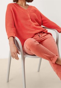 Maglione a maglia color corallo con scollatura a V e texture a costine, abbinato a jeans slim-fit color corallo. Sedia grigia moderna in un ambiente neutro.