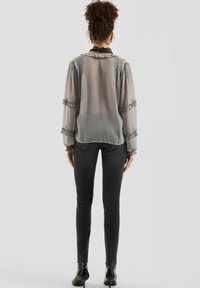 Przezroczysta szara bluzka z falbankami na rękawach i kołnierzyku, w połączeniu z czarnymi obcisłymi dżinsami i botkami, widziana od tyłu.