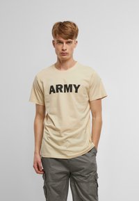 Bēšas kokvilnas t-krekls ar īsām piedurknēm un apaļu apkakli, ar izteiksmīgu melnu "ARMY" tekstu pāri krūtīm. Valkāts ar pelēkiem kuplajiem biksēm.