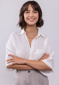 Witte korte mouwen knoopshirt gecombineerd met lichtgrijze hooggelegen broek. Een delicate gouden ketting met groene accenten.