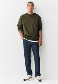 Olivgrön sweatshirt med ribbade muddar, bärs över en vit skjorta. Ett par mörkblå denimjeans och vita sneakers med grå detaljer.