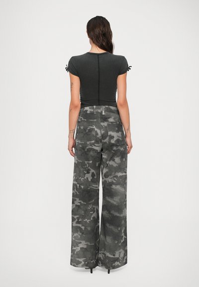Femme aux longs cheveux bruns, portant un t-shirt noir ajusté et un pantalon large en camouflage gris taille haute, se tenant de dos contre un fond blanc.