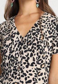 Blusa con stampa leopardata caratterizzata da scollatura a V, maniche corte e bottoni scuri, realizzata in un tessuto testurizzato con diverse tonalità di beige e nero.