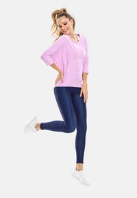 Hellrosa, lockeres Oberteil mit 3/4 Ärmeln; kombiniert mit dunkelblauen, glänzenden Leggings. Das Model trägt weiße Sneakers und posiert dynamisch.