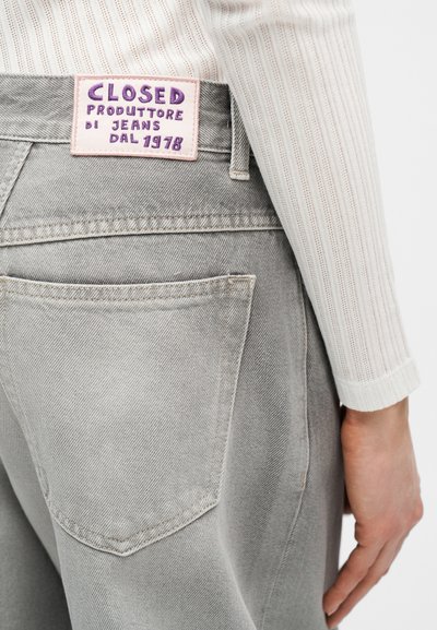 Κοντινό πλάνο στην πίσω τσέπη και τη μέση γκρι τζιν με ένα ροζ ετικέτα που γράφει «Closed Produttore di Jeans dal 1978», φορεμένα με ένα λευκό ριμπ ύφασμα μανίκι.