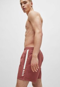 Røde badeshorts lavet af let materiale, med en elastisk talje, hvide sideaccenter og et logo vist vertikalt.