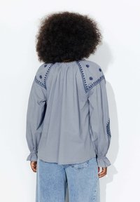 Blouse grise brodée avec des manches longues et un col froncé. Présente des broderies florales bleues sur les épaules et des poignets élastiques. Coupe classique.