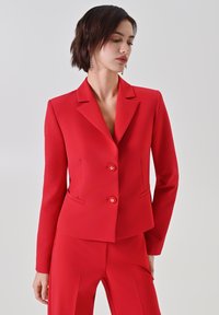 Giacca blazer rossa con taglio sartoriale, rever a lancia, due bottoni frontali e tasche laterali. Tessuto liscio con silhouette strutturata.