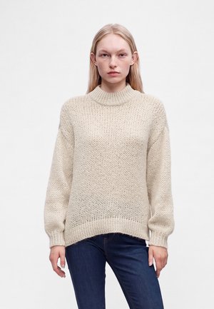 Jeune femme aux cheveux blonds raides portant un pull en tricot beige et un jean bleu foncé, debout devant un fond blanc uni.