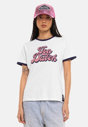 Jeune femme portant un t-shirt blanc Von Dutch avec un liseré bleu marine et une casquette rose Von Dutch, debout avec une expression neutre.