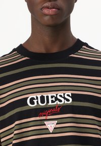 Svart, grønn og rosa stripete t-skjorte med rund hals. Har hvit og rød "GUESS" logo og trekantet merke nederst.