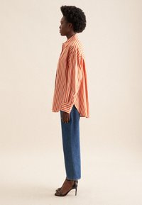 Camicia oversized a strisce arancione e bianca con maniche lunghe, abbinata a jeans blu a vita alta e sandali neri con tacco. Texture liscia.