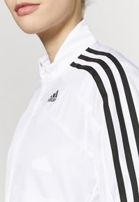 Chaqueta blanca de adidas con cuello alto, diseño de tres franjas negras en los hombros y un acabado de tejido texturizado.