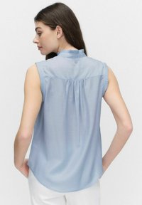 Lysblå ærmeløs bluse med en flæsekrave, knapdetaljer bagpå, og samlet stof langs skulderstykket for ekstra tekstur.