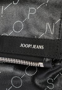 Borsa nera texture con lettere e cerchi bianchi stampati, dotata di chiusura con zip e un'etichetta in tessuto nera con la scritta "JOOP! JEANS."