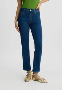 Jean en denim taille haute de couleur bleue, comportant cinq poches, une jambe droite slim et de subtils détails de couture dorée.