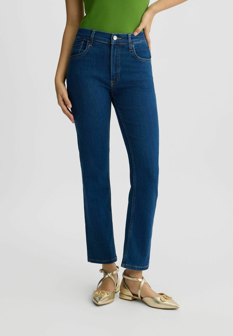 Jean en denim taille haute de couleur bleue, comportant cinq poches, une jambe droite slim et de subtils détails de couture dorée.
