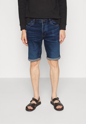 Dunkelblaue Denim-Shorts im entspannten Schnitt, mit umgeschlagenen Säumen und sichtbaren Nähdetails. Kombiniert mit schwarzen Sandalen, die den Komfort betonen.
